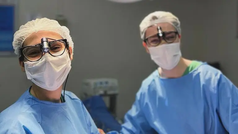 Dr. Lucas Cunha e Dra. Izadora Reis com roupas cirúrgicas, realizando cirurgia plástica com olhar alegre.